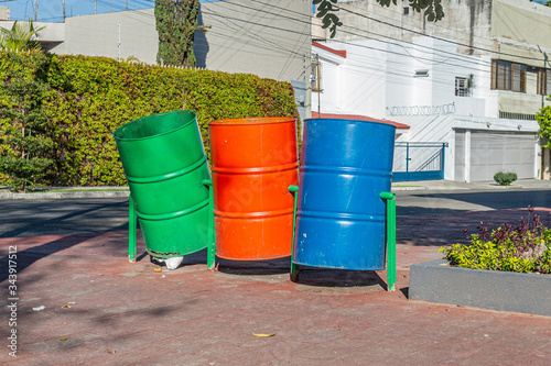 Three rotating empty metal barrel trash bins on a metal base on a sidewalk in a city public garden with houses in the background, sunny day in Guadalajara, Jalisco, Mexico © Emile