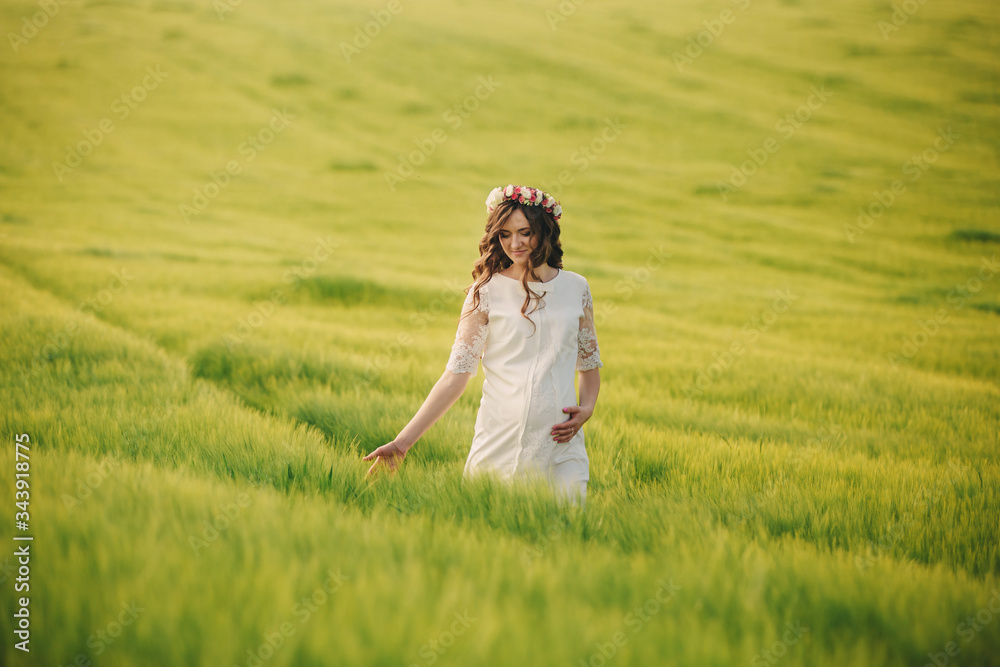 pregnant woman in white dress with wreath of flowers on head is relaxing outdoors in grass field. motherhood concept, selective focus