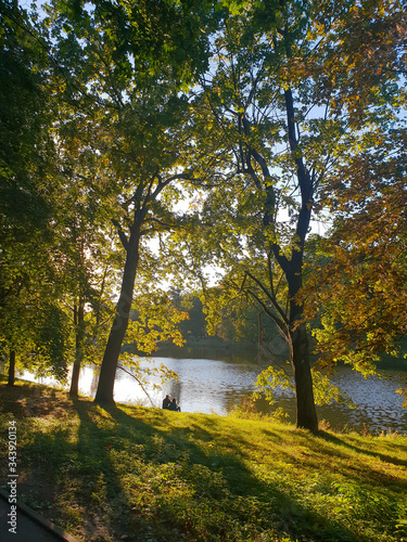 Para siedząca nad brzegiem stawu pomiędzy drzewami w słoneczny jesienny dzień w parku im. Mickiewicza w Łodzi.