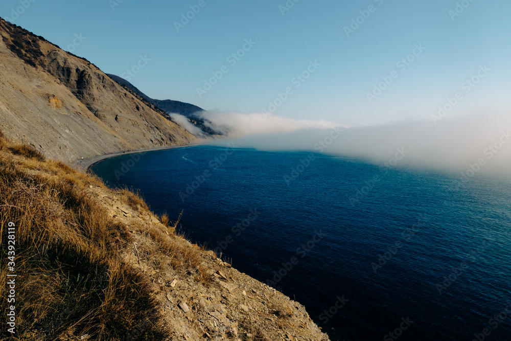 The edge of the juniper forest of Utrish Nature Reserve over the blue sea over which dense fog spreads and finds on high coastal mountains.