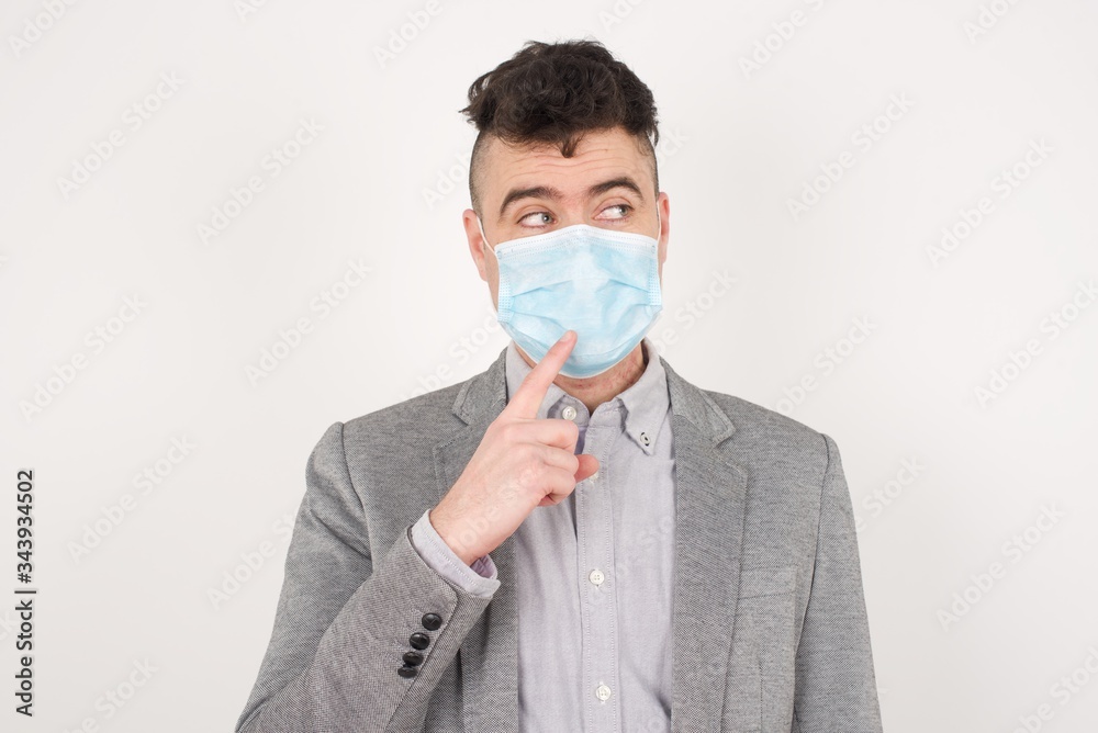 Dreamy male with thoughtful expression, looks to the camera, keeps hand near face, bitting a finger thinks about something pleasant, poses against gray wall. Dreamy male with thoughtful expression, looks to the camera, keeps hand near face, bitting a finger thinks about something pleasant, poses against gray wall.