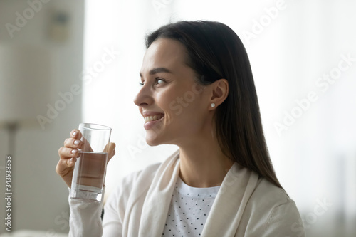 Smiling young Caucasian woman look in distance drinking clean pure mineral water at home, happy millennial female hold glass enjoy clear still aqua for body refreshment, healthy lifestyle concept