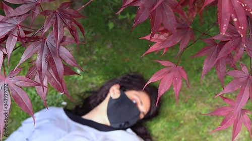 red maple leaves in the wind and blurred brunette girl wearing black medical mask laying on the grass, concept of life during coronavirus pandemic, new normal