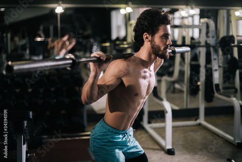 Slim bearded young man with muscular wiry naked torso performing barbell squats during sport workout training in modern dark gym. Concept of healthy lifestyle.