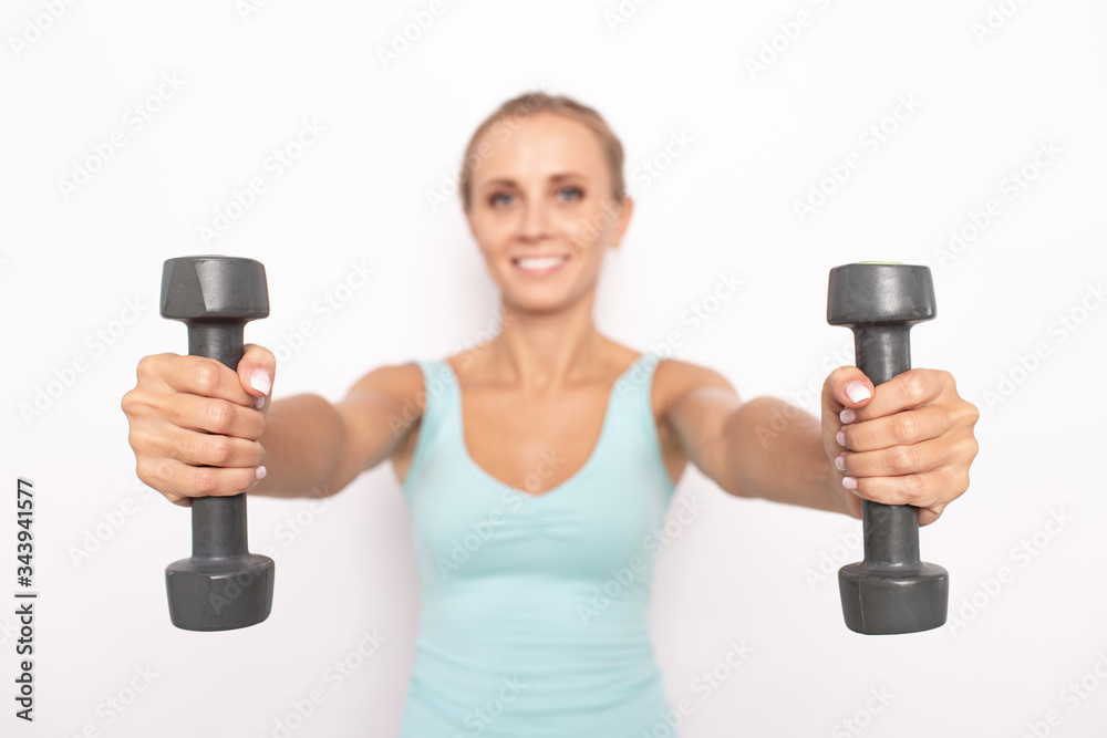 Smiling blond woman in blue tip holds two dumbbells in hands. Selective focus. White isolated background Smiling blond woman in blue tip holds two dumbbells in hands. Selective focus. White isolated background