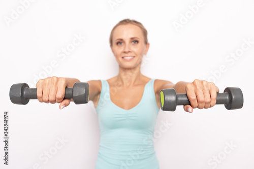 Smiling blond woman in blue tip holds two dumbbells in hands. Selective focus. White isolated background