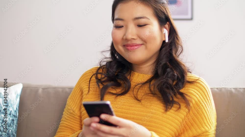 people, technology and leisure concept - happy asian young woman with wireless earphones and smartphone listening to music at home people, technology and leisure concept - happy asian young woman with wireless earphones and smartphone listening to music at home
