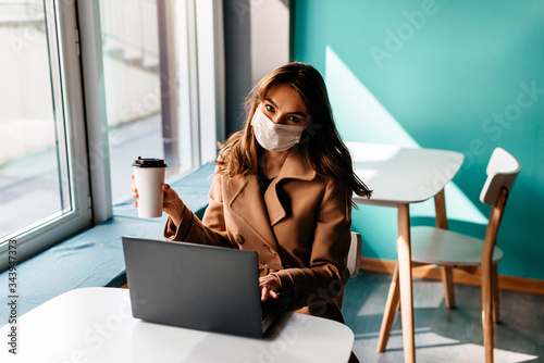 Beautiful woman in a protective mask sits in a cafe at a laptop. Protecting the population from viruses