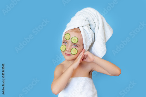 A pretty little girl in a towel on her head, makes a mask out of cucumbers and smiles on a blue background