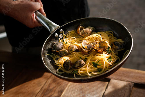 padella con spaghetti alle arselle tenuta da una mano  , sullo sfondo  tavolo di legno photo