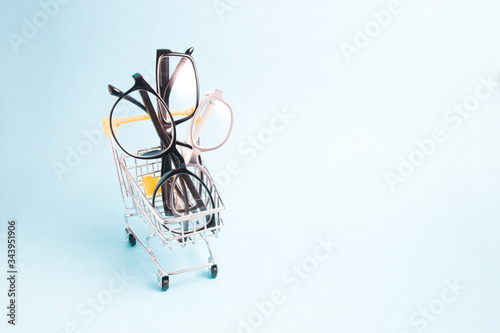 children's glasses and two pairs of black glasses for adults in a small shopping cart, blue background, copy space, online purchase of glasses and eyeglass frames