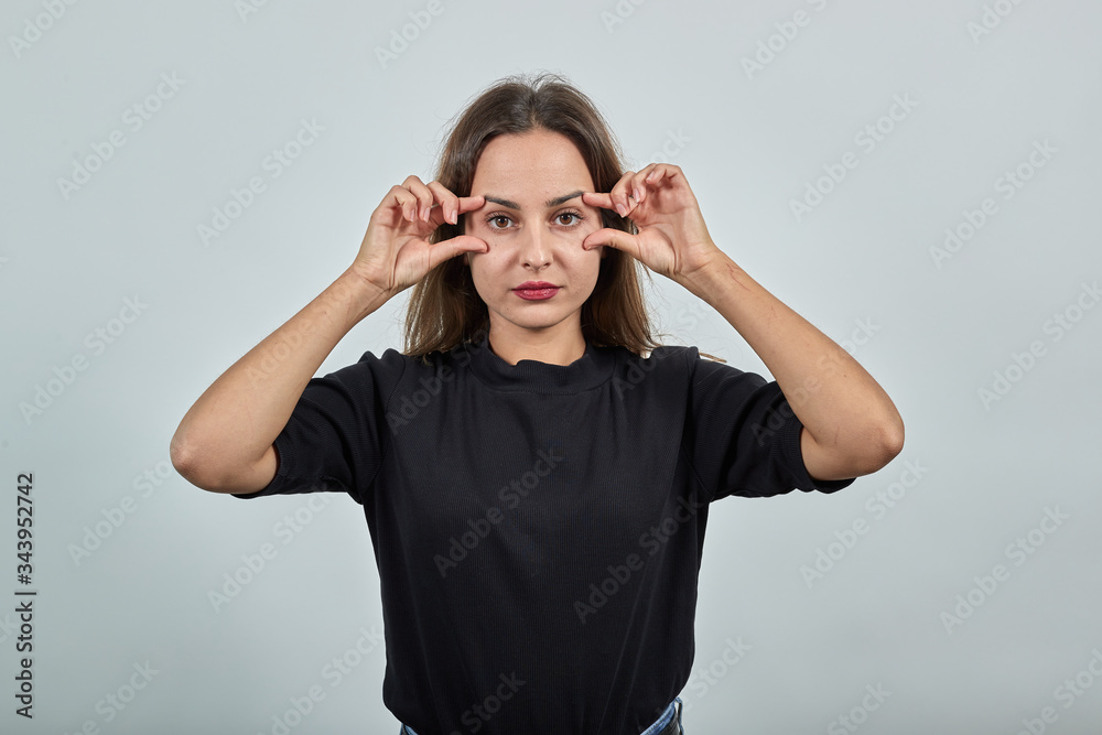 Cute young brunette woman in black t-shirt, blue jeans with belt on gray background, tired girl opened her eyes with fingers Cute young brunette woman in black t-shirt, blue jeans with belt on gray background, tired girl opened her eyes with fingers