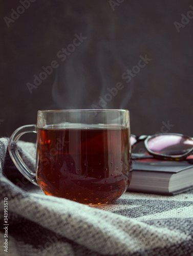 Steam over a transparent cup of black tea that stands on a gray plaid. Over a cup of tea are a book and glasses