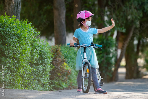 Fototapeta Naklejka Na Ścianę i Meble -  Cute little girl with mask on face ridding a bike and salute other kids from distance.