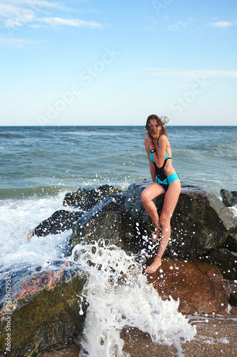 A young girl sits on stones at the sea, splashing waves, waves are breaking on stones, emotions of delight and happiness. © Ruslan