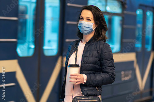 Beautiful girl, young woman standing at station in underground, metro, subway, waiting for public transport, train in medical protective mask on her face. Virus pandemic coronavirus concept. Covid-19