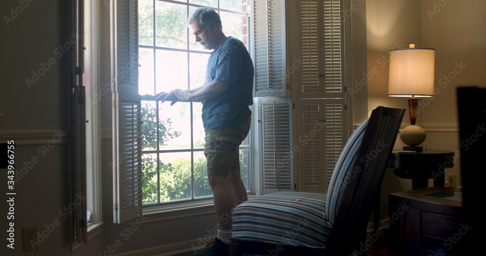 A depressed mature man standing at his window feeling isolated and fearful during the coronavirus pandemic business closures as well as market woes.