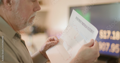 Canvas Print A mature man looks at his investment statement containing nothing but bad news because of COVID19 pandemic with stock market graphs and charts on a TV in the background