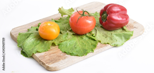  Culinary composition of fresh natural vegetables on a white background: red, yellow tomatoes, bell peppers and green fresh salad lie on a wooden cutting board