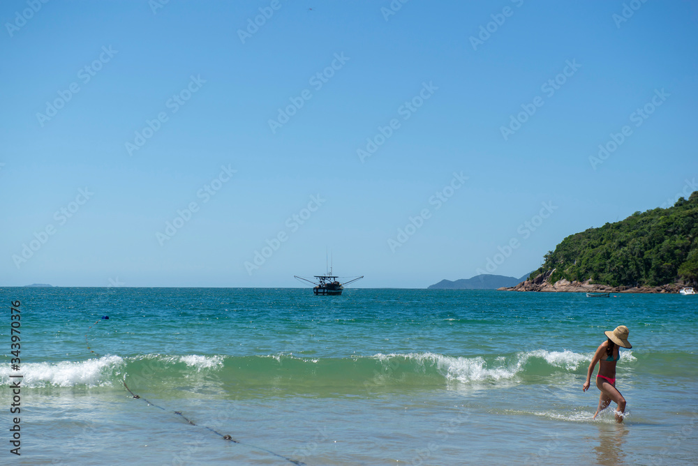 View of a beautiful beach in Brazil with coast with beautiful trees, turquoise sea and clear sky. Unrecognizable woman in bikini with hat coming out of the sea. Space for text and advertisement.
