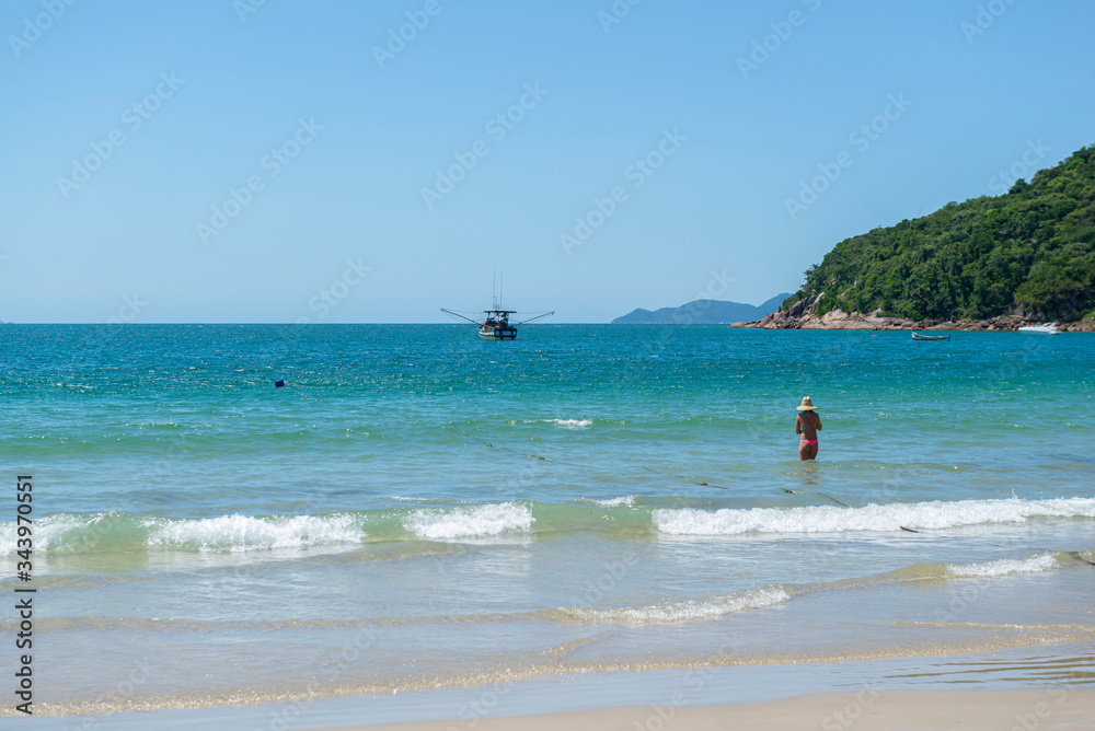 View of a beautiful beach in Florianópolis - Brazil with coast with beautiful trees, turquoise sea and clear sky. Unrecognizable woman in bikini with hat in the sea. Space for text and advertisement.