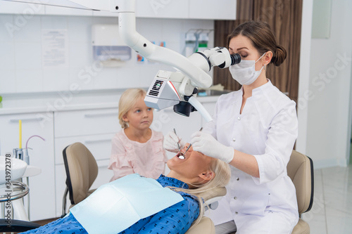 A dentist examines the oral cavity of a woman with the help of a special medical appliance  while her little daughter is watching them