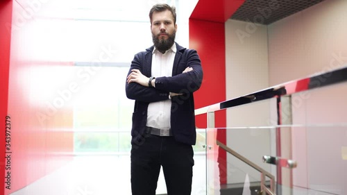 Confident bearded man is standing in front of the camera near glass fence in modern building crossing his hands. Close-up view of serious brunette guy with folded arms in business center with red