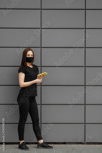 Young beautiful woman wear black face mask protection against epidemic coronavirus covid-19. Looks on phone screen. European girl medical protective mask orange business suit
