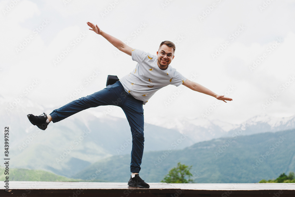 A young cheerful guy makes a face against the background of mountains in full height and stands on one leg, raising his hands up. A young cheerful guy makes a face against the background of mountains in full height and stands on one leg, raising his hands up.