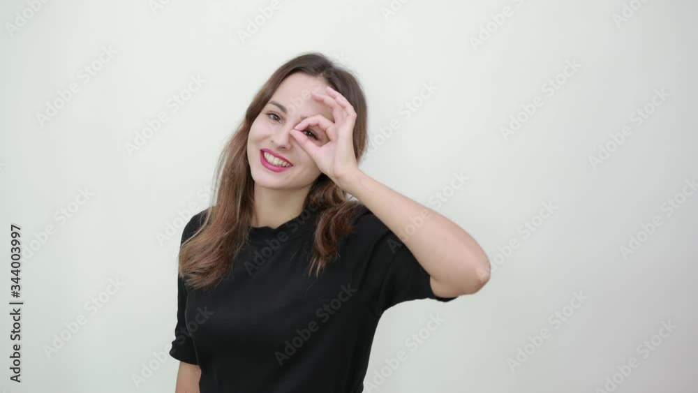 Pretty young brunette in a black t-shirt, blue jeans with a belt on gray background, happy smiling girl holds an OK sign near her eye, pointing with hand