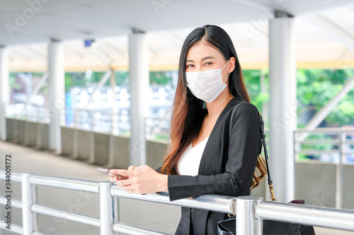 Portrait of business woman with hygiene mask stand along walk way and hold mobile phone, she also look forward and look relax during covid-19 pandemic in the city.