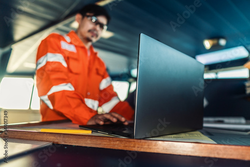 Filipino deck Officer on bridge of vessel or ship wearing coverall during navigaton watch at sea . He is using laptop, electronic paperwork at sea, concept of reporting