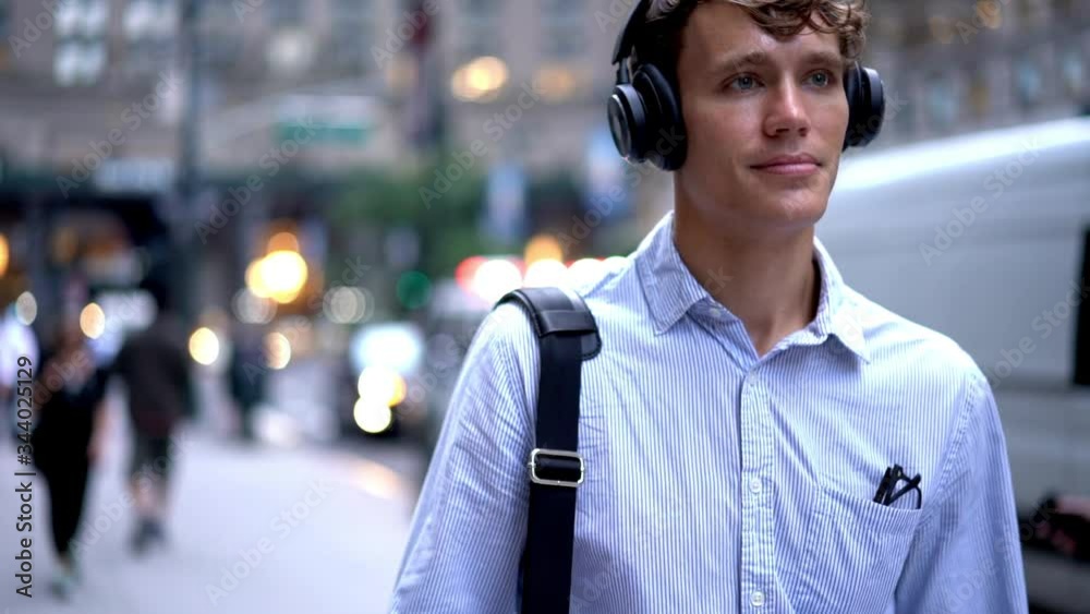 Young professional manager in electronic digital heaphones walking at city street and enjoying way time for listening music set during multitasking, slow motion of employee in modern earphones
