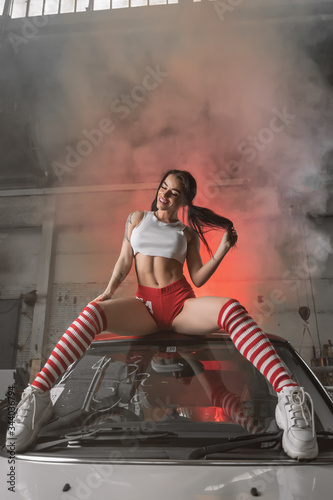 Attractive Caucasian female in top and shorts sitting on roof silver car in autostation against white smoke