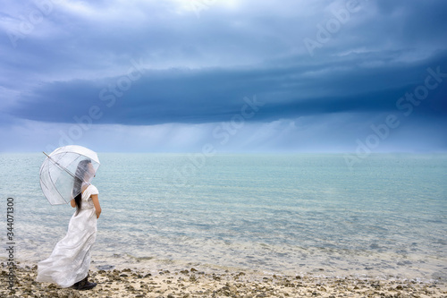 Woman wearing white dress holding umbrella standing on the beach over sea water and storm clouds sky background, Bahrain.