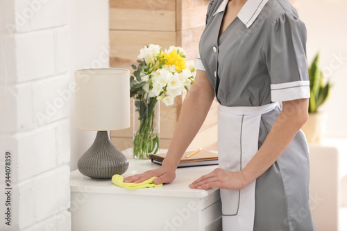 Young chambermaid wiping dust from furniture in hotel room, closeup photo