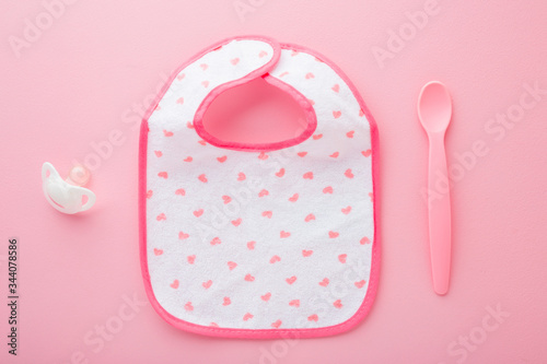 Foto Baby textile bib, plastic spoon and soother on light pink table background