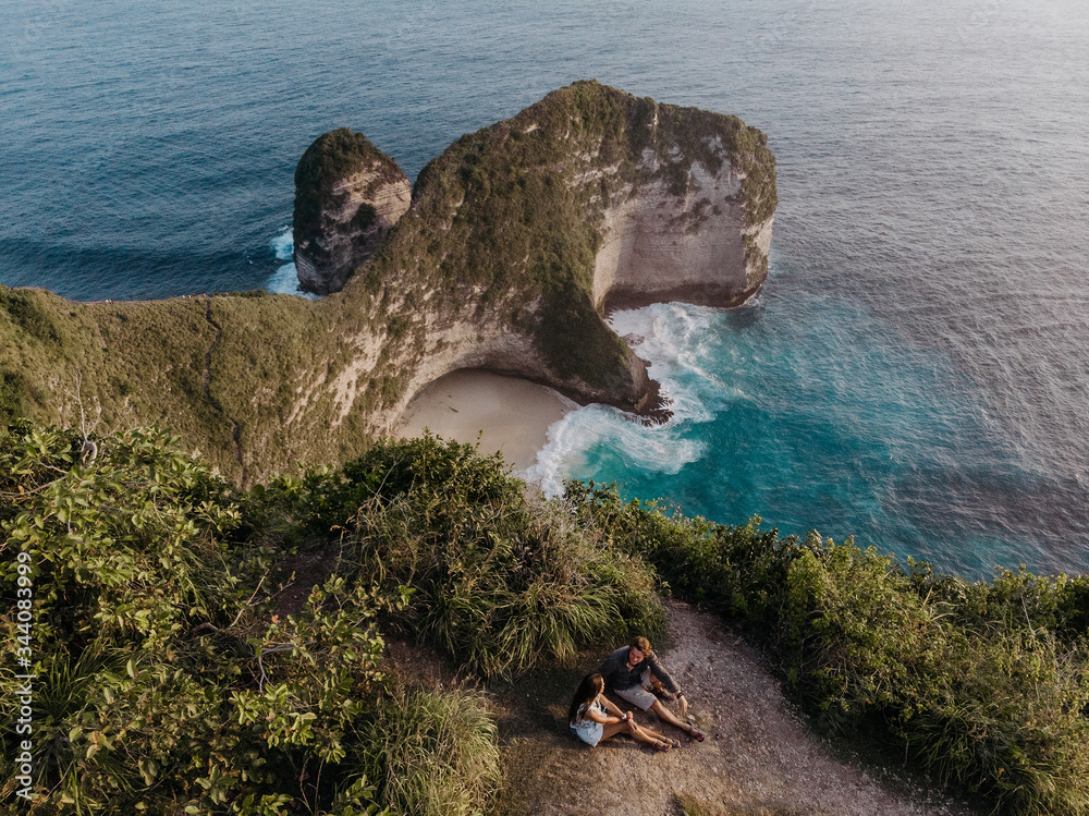 Couple sitting on the clif and looking on popular place in Nusa Penida - Kelingking beach. Travelling couple. Aerial view of the small island of Nusa Penida Island