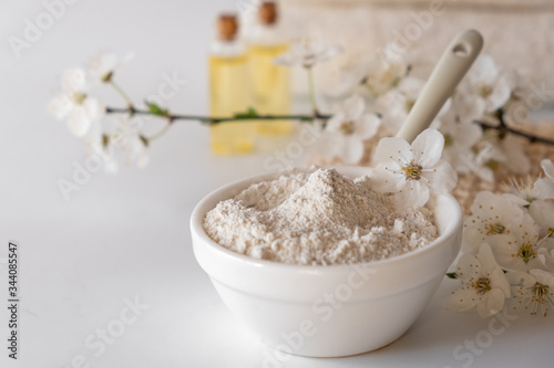 Ceramic bowl with white clay powder on white background. Ingredients for homemade facial and body mask or scrub and fresh sprig of flowering cherry. Spa and bodycare concept.