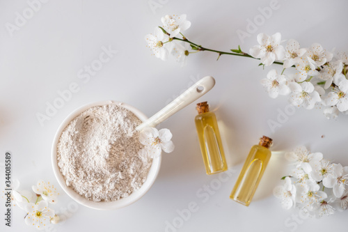 Ceramic bowl with white clay powder on white background. Ingredients for homemade facial and body mask or scrub and fresh sprig of flowering cherry. Spa and bodycare concept.