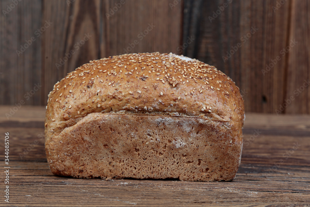 Side view of fresh bread sprinkled with sesame seeds. A freshly baked molded, golden loaf on a wooden, brown, rustic table. In the background brown boards.