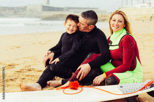 Happy family with surfboard on ocean coast. Cheerful parents and cute little son in wetsuits sitting together on sandy coast. Water sport concept