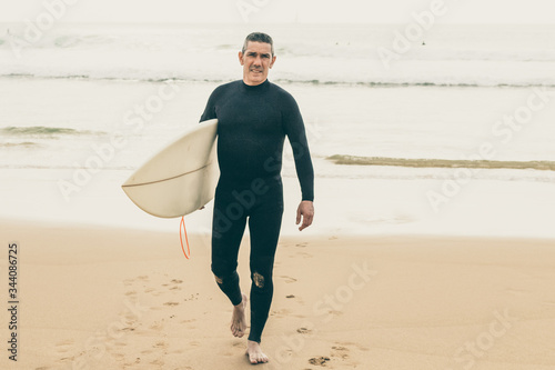Handsome male surfer looking at camera. Full length view of middle aged man in wetsuit holding surfboard and walking on sandy sea coast. Surfing concept