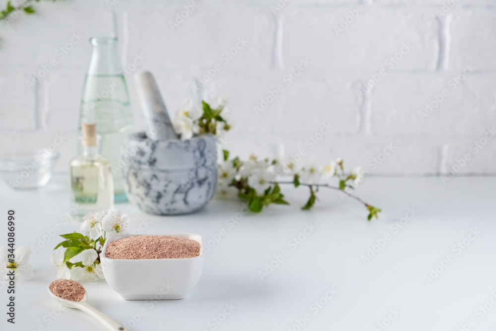 Ceramic bowl with red clay powder, ingredients for homemade facial and body mask or scrub and fresh sprig of flowering cherry on white background. Spa and bodycare concept.