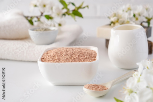Ceramic bowl with red clay powder, ingredients for homemade facial and body mask or scrub and fresh sprig of flowering cherry on white background. Spa and bodycare concept.