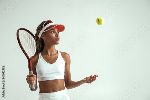 Ready to play. Portrait of young african woman in sports clothes holding tennis racket on her shoulder and throwing tennis ball while standing against grey background © Friends Stock