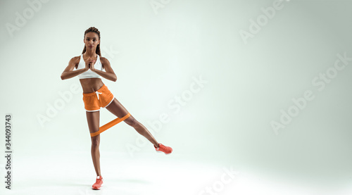 Exercises for perfect legs. Full length of young and slim african woman in sports clothing exercising with a resistance band and looking at camera while standing in studio against grey background