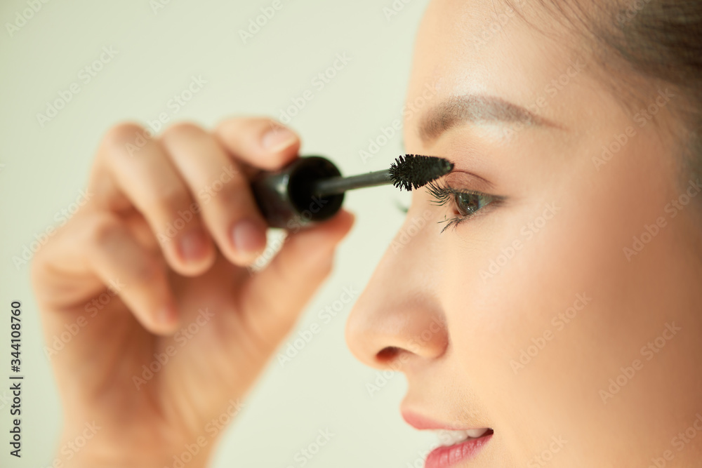 Portrait of young Asian woman applying mascara over light background.
