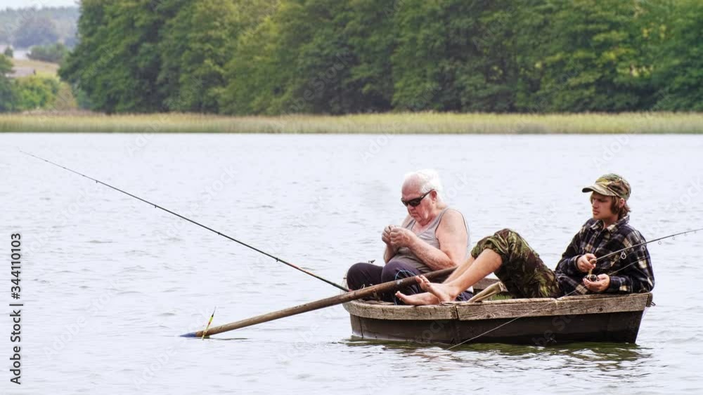 Old angler with a grandson sitting in the boat on the lake with a rod while fishing. Outside. Old angler with a grandson sitting in the boat on the lake with a rod while fishing. Outside.