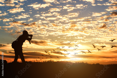 Silhouette of male photographer taking picture against sunset with a flock of birds flying.